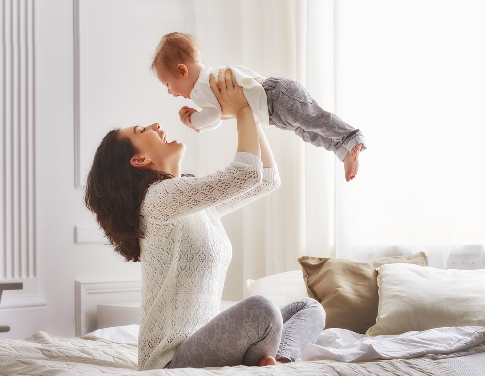 Happy mother plays with her baby on her bed after restoring her pre-pregnancy body.