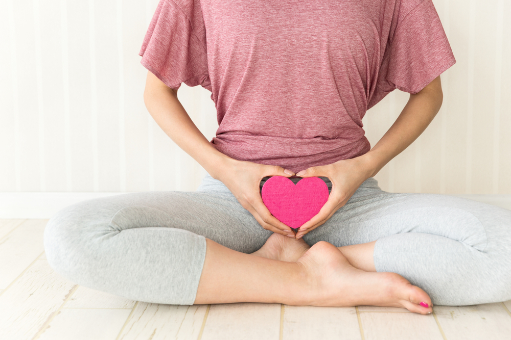 Woman in exercise attire holds a heart figure in front of her vagina.